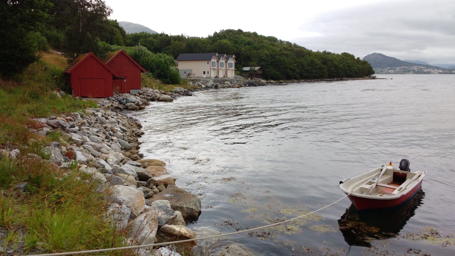 Røde naust ved fjorden med motorbåt, grønne lier og fjell i bakgrunnen -- utsikten fra Martingarden
